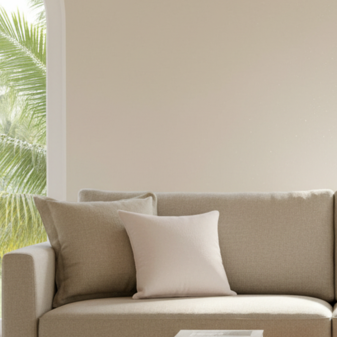 Beige sofa with two pillows in a room with a light-colored wall and greenery.