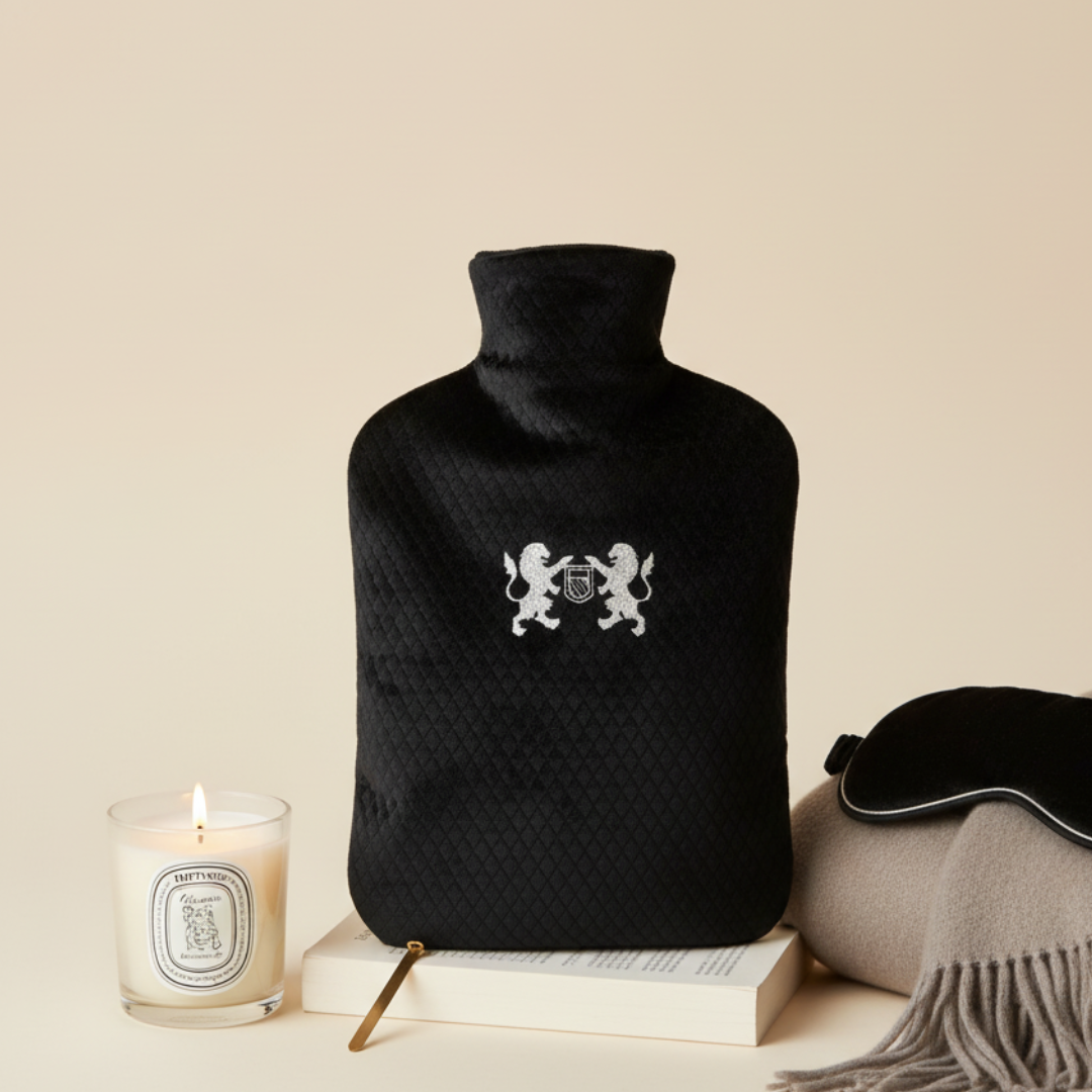 Black hot water bottle with quilted texture, Diptyque candle, and eye mask on a beige background