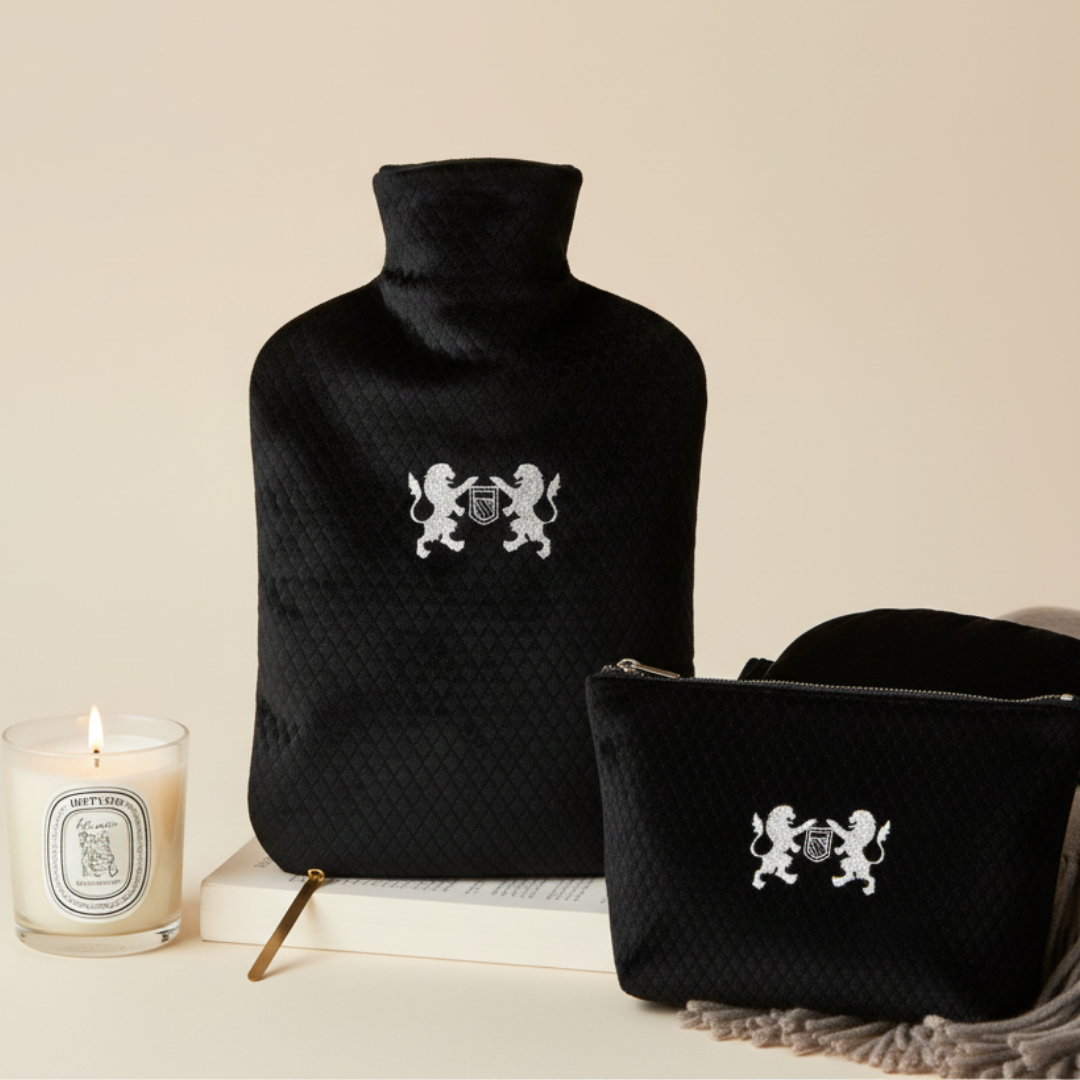 Black quilted hot water bottle with decorative emblem, matching pouch, and Diptyque candle on a beige background
