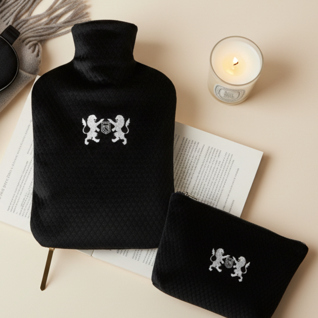 Black hot water bottle with quilted cover and emblem on a beige surface with a lit candle.